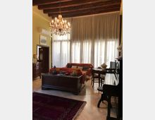 Soggiorno in stile classico con soffitto a travi in legno, grande lampadario a bracci e ampie finestre con tende bianche. Nella stanza ci sono divani, un tavolo basso centrale, un pianoforte lungo la parete e un piccolo tavolo con sedie vicino alle finestre.