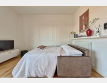 Camera da letto moderna con pareti bianche a riquadri e pavimento in legno, con un divano-letto rivestito in tessuto e biancheria chiara. A lato ci sono un mobile basso con TV e, sopra un termosifone, una mensola con libri, lampada, vaso rosso e una stampa incorniciata.