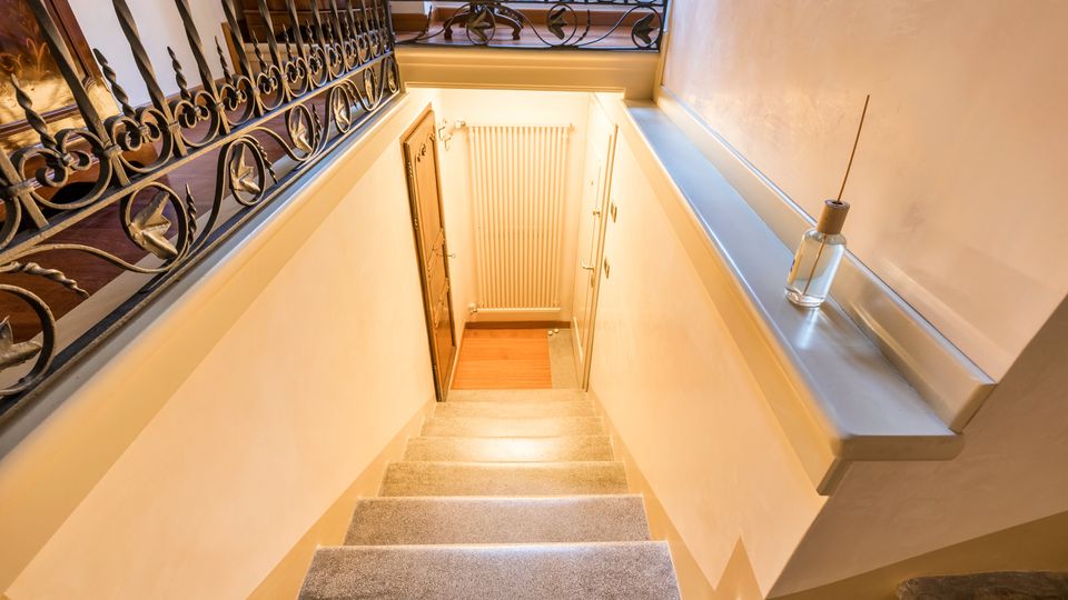Vano scala interno con gradini in pietra che scendono verso un pianerottolo in legno, illuminato da luce calda. Ringhiere in ferro battuto decorato lungo il parapetto e pareti chiare con termosifone sul fondo.