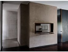 Interno di un’abitazione con grande volume murario intonacato al centro, attraversato da una finestra passavivande con ripiano in legno che affaccia su una cucina con mobili bianchi. A destra si vedono alte ante scure e a sinistra un corridoio con pareti chiare e pavimento in legno scuro.