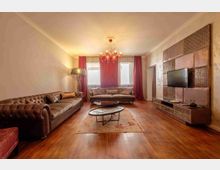 Soggiorno moderno con pavimento in legno, due divani in pelle e tavolini ovali su un tappeto centrale. Sul fondo ci sono grandi finestre con tende rosse e un lampadario, mentre a destra una parete rivestita a pannelli ospita una TV sopra un mobile basso.