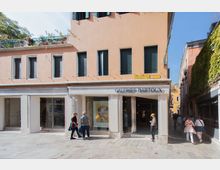 Facciata di un edificio color pesca con portico e grandi vetrine d’ingresso della galleria d’arte “Galeries Bartoux”, su una piazza pavimentata in pietra. A destra si apre una stretta via tra edifici, con un muro ricoperto di edera e segnaletica stradale sopra l’angolo dell’edificio.