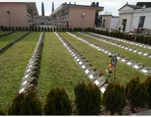 Cimitero con file ordinate di tombe a prato, delimitate da pietre inclinate con targhette metalliche, e siepi di piccoli cipressi lungo i bordi. Sullo sfondo si vedono strutture con loculi e una cappella bianca con croce.