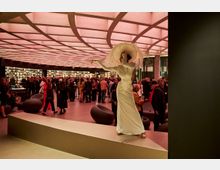 Spazio interno moderno con soffitto a griglia retroilluminato di colore rosa, arredi minimalisti e tavoli circondati da scaffali pieni di oggetti espositivi. Nell'area si trovano persone in piedi e sedute, in un contesto che suggerisce un evento o una mostra.