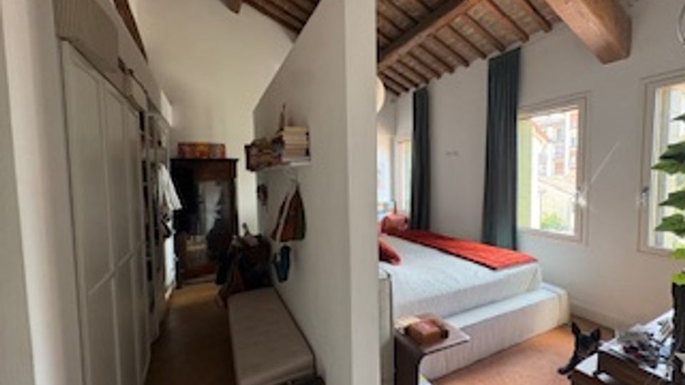 Interno di una camera con soffitto alto a travi di legno e pavimento in legno, divisa da una parete: da un lato armadi e panca lungo il corridoio, dall’altro una zona letto con finestra e tende scure. Arredi moderni essenziali e luce naturale proveniente dalle finestre.