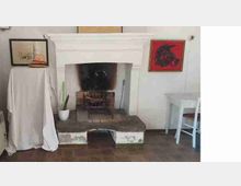 Caminetto in muratura bianca con ampia bocca e focolare annerito, su base in pietra, in una stanza con pavimento in legno. Ai lati ci sono quadri alle pareti, una pianta in vaso, un mobile coperto da un telo e un piccolo tavolo con sgabello.