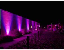 Esterno notturno di un lungo edificio moderno con parete in pannelli orizzontali, illuminata da faretti viola che creano fasci verticali. In primo piano si vede un sentiero tra sabbia e paletti con corde, con strutture tipo gazebo sulla destra.