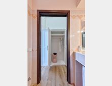 Vista attraverso una porta con cornice in legno che collega un bagno piastrellato in rosa a un piccolo spazio con pavimento in legno e un armadio bianco aperto con ripiani e gruccia. A destra si vede un piano lavabo con specchio e accessori.