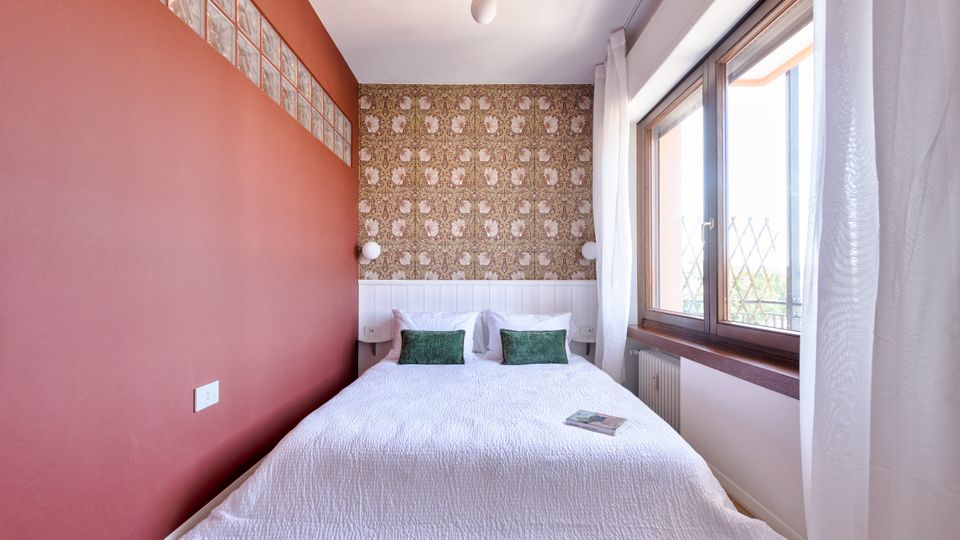 Camera da letto stretta con letto matrimoniale rivestito di coperta bianca e due cuscini verdi, parete laterale color terracotta e parete di fondo con carta da parati decorata e boiserie bianca. A destra, grande finestra con telaio in legno e tende bianche che lascia entrare molta luce naturale.