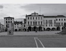 Ampia piazza lastricata con edifici storici a più piani sullo sfondo, con portici ad arcate e una facciata centrale decorata sormontata da un orologio. In primo piano si vedono il selciato, un grande pannello informativo e parte di un monumento in pietra ai margini della piazza.