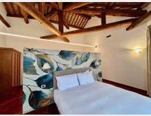 Camera da letto con soffitto alto e travi in legno a vista, pareti chiare e pavimento in legno. Letto matrimoniale con testiera imbottita davanti a una grande carta da parati con foglie blu, applique ai lati e armadio in legno.