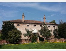 Edificio storico in mattoni con tetto in coppi e due alte canne fumarie, con una fila di piccole finestre ovali lungo la facciata. Davanti si vede un prato verde con fiori gialli, un muretto in mattoni e alberi e arbusti che parzialmente coprono l’ingresso con una piccola copertura arcuata.