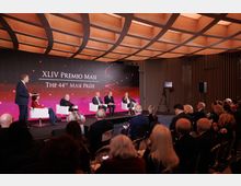 Evento in una sala conferenze moderna con soffitto in legno a griglia e pareti con rivestimento verticale a doghe. Sul palco, uno sfondo luminoso con la scritta "XLIV Premio Masi - The 44th Masi Prize" e diverse persone sedute su poltrone bianche.