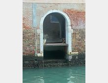 Ingresso ad arco in un edificio storico in mattoni a Venezia, che si affaccia direttamente su un canale con acqua verde; la base dell'entrata mostra segni di erosione causati dall'acqua.