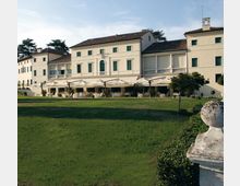 Grande edificio in stile villa storica color crema con persiane verdi e tetto in coppi, affacciato su un ampio prato. Davanti alla facciata ci sono una terrazza con tende bianche e un portico con colonne, circondati da alberi e siepi.