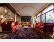 Ampio salone arredato in stile classico con grandi divani rossi, tappeto persiano e tavolino centrale su pavimento a scacchi. Lungo un lato si aprono alte finestre ad arco con vetrate a reticolo, con travi in legno a vista e un grande dipinto murale sulla parete di fondo.