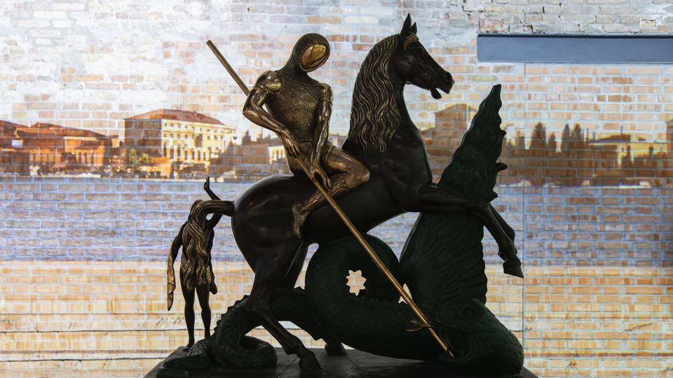Scultura in bronzo di un cavaliere a cavallo con lancia che combatte un drago, esposta su una base in una sala interna. Sullo sfondo si vede una parete di mattoni chiari con una proiezione panoramica di un paesaggio urbano.