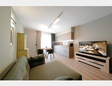 Soggiorno open space moderno con divano in primo piano, tavolo da pranzo vicino a una grande finestra con tende e cucina lineare con pensili e illuminazione sottopensile. A destra c’è un mobile basso con televisore, pavimento in legno chiaro e illuminazione a binario sul soffitto.