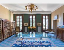 Salotto in stile classico con tre alte finestre a vetri e telai in legno su parete verde, radiatori sotto le finestre e lampadario decorato al soffitto. Al centro c’è un tavolo basso in vetro su tappeto geometricamente decorato, con mobili in legno e sedute lungo le pareti.