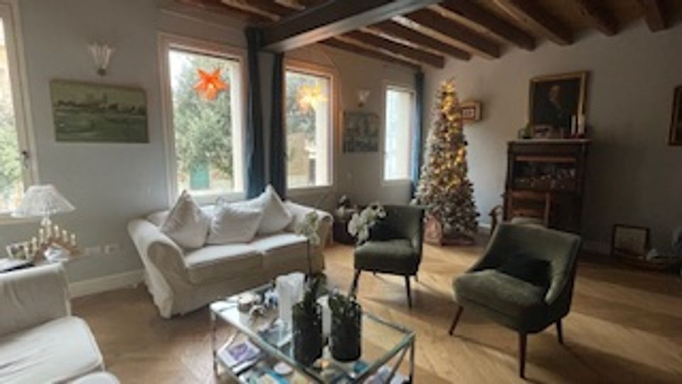 Soggiorno rustico con travi in legno a vista, divano chiaro e due poltrone scure attorno a un tavolino in vetro. Sullo sfondo si vedono una finestra con luce naturale, un camino e un albero di Natale decorato vicino alla parete.