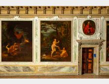 Parete interna di una sala storica decorata con grandi dipinti incorniciati e pannelli a finto marmo nella parte inferiore. A destra si vede una porta lignea scura inserita in una cornice architettonica scolpita, con un piccolo ritratto ovale sopra l’architrave.