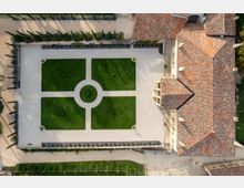 Veduta aerea dall’alto di un giardino formale rettangolare con quattro aiuole di prato divise da vialetti chiari e un elemento circolare centrale. A destra si affianca un grande edificio con tetto in tegole e, lungo il perimetro, filari di alberi e siepi delimitano i camminamenti.