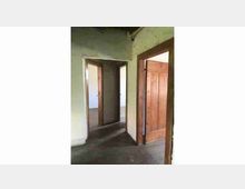 Interno di un edificio con pareti intonacate chiare e pavimento scuro, con più porte in legno aperte che conducono a stanze adiacenti. Gli stipiti e i bordi delle aperture risultano semplici e la luce entra dalle stanze sullo sfondo.