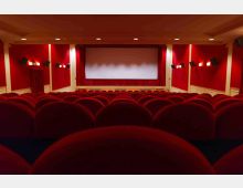 Interno di una sala cinematografica con file di poltrone rosse rivolte verso un grande schermo bianco sul palco. Pareti rosse con pannelli chiari, luci a parete e faretti a soffitto illuminano l’ambiente.