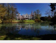 Veduta di una grande villa in stile neogotico con torrette e facciata chiara, situata su un prato e circondata da alberi. In primo piano un lago calmo riflette l’edificio e la vegetazione sotto un cielo sereno.