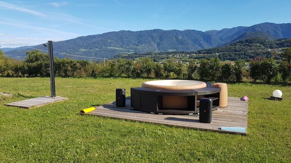 Vasca idromassaggio rotonda su una pedana in legno in un prato aperto, con un piccolo punto doccia esterno nelle vicinanze. Sullo sfondo si vedono colline e montagne boscose sotto un cielo sereno.