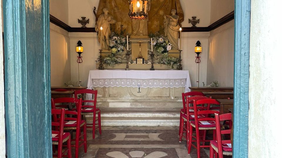 Interno di una piccola cappella con altare centrale decorato da statue e fiori, illuminato da una lampada sospesa e applique laterali. Il pavimento in marmo presenta un motivo geometrico con rosetta centrale e ai lati ci sono file di sedie rosse, visto dalla soglia di una porta azzurra.