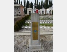 Cimitero con un cippo commemorativo in pietra con croce e targa metallica in primo piano, circondato da vialetti in ghiaia. Sullo sfondo si vedono file di lapidi a terra, piccoli mausolei bianchi e alti cipressi.