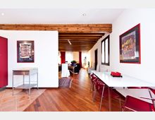 Interno di un appartamento con ambiente open space e soffitto con travi in legno a vista, pavimento in parquet e pareti bianche. A destra c’è un tavolo moderno con sedie rosse e finestre, mentre sul fondo si vede la zona soggiorno con tende rosse e arredi.