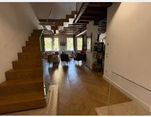 Interno di un’abitazione moderna con scala in legno a vista e pavimento in parquet disposto a spina di pesce. Ambiente open space con soffitto con travi in legno, zona pranzo e ampie vetrate sul fondo che affacciano sul verde esterno.