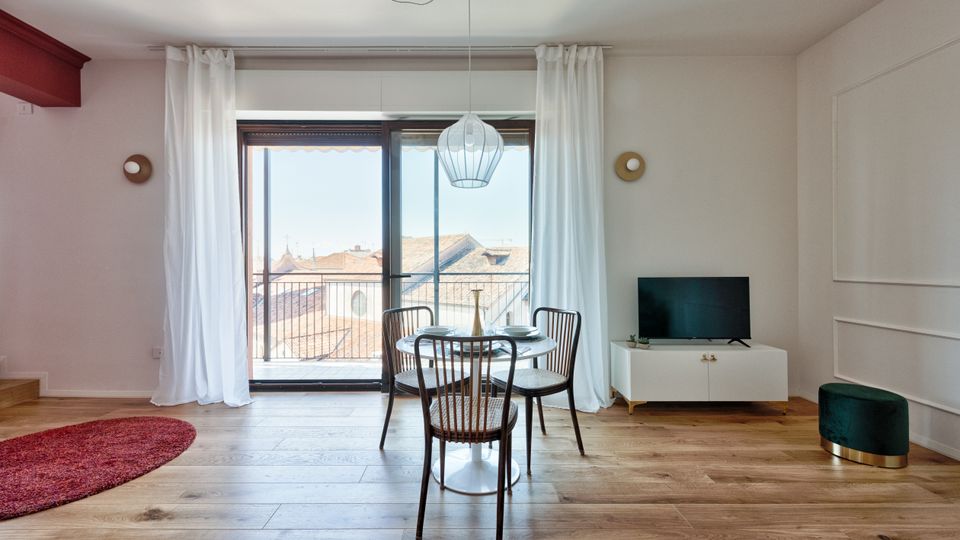 Soggiorno moderno con pavimento in legno, tavolo rotondo con quattro sedie e lampada a sospensione, affacciato su una grande porta-finestra con tende bianche che dà su un balcone con vista sui tetti. A destra ci sono un mobile basso con televisore e un piccolo pouf, con pareti bianche decorate da applique rotonde.