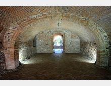 Interno di una cantina con volte a botte in mattoni e pareti in pietra grezza, con un pavimento in cotto disposto a spina di pesce. Sullo sfondo, una porta ad arco con tende leggere lascia intravedere un giardino illuminato.