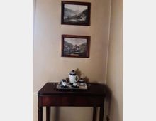 Angolo di una stanza con pareti beige, un tavolino in legno scuro con un vassoio e servizio da tè in ceramica bianca e blu. Sopra il tavolino sono appesi due quadri incorniciati con paesaggi montani.