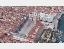 Veduta aerea di una grande piazza lastricata circondata da edifici storici con portici, dominata da un alto campanile e da una basilica con cupole. Sul lato destro si vede il lungomare con banchine e barche, mentre intorno si estende un fitto tessuto urbano di case con tetti in terracotta.