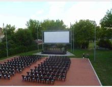 Arena cinematografica all’aperto in un parco, con file di sedie nere disposte su una pavimentazione rossa di fronte a un grande schermo bianco su palco. Intorno ci sono alberi e prato, con alcuni lampioni e edifici visibili sullo sfondo.