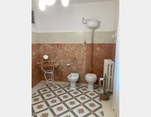 Bagno in stile vintage con pavimento a mosaico geometrico a stelle e pareti rivestite in marmo rosato e beige. Sono visibili bidet e WC con cassetta alta a tirante, un piccolo tavolino in ferro e un termosifone a colonna.