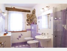 Bagno luminoso con piastrelle bianche e viola, pavimento a scacchi e finestra con tenda chiara e trave in legno sopra. A destra ci sono lavabo con specchio, bidet e WC, e una doccia con box in vetro accanto a una parete con inserto in pietra.