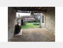 Portico rustico con pavimento in ghiaia e soffitto a travi in legno, che si apre su un cortile erboso. Sullo sfondo si vedono edifici rurali in muratura con tetti in coppi e un piccolo capanno.