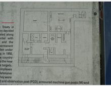 Pannello informativo con pianta tecnica di un bunker o struttura militare sotterranea, con stanze etichettate (camera, sala comando, docce, deposito munizioni, WC) e corridoi. A sinistra si intravede testo esplicativo in inglese e a destra il bordo metallico della cornice del pannello.
