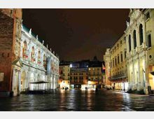 Piazza cittadina di notte con ampia pavimentazione in pietra lucida, fiancheggiata da palazzi storici illuminati. A sinistra un edificio con portico ad arcate e statue sul cornicione, a destra una facciata con logge e archi al piano superiore, con negozi sotto i portici.