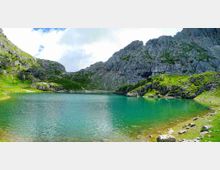 Lago alpino dalle acque turchesi circondato da pendii erbosi e rocce, con una riva sassosa in primo piano. Sullo sfondo si alzano alte pareti montuose grigie sotto un cielo parzialmente nuvoloso.