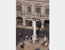 Veduta dall’alto di una piazza lastricata con una colonna monumentale in pietra su basamento, sormontata da una statua. Sullo sfondo si vede la facciata di un edificio storico con portici ad archi, balconcini e finestre con persiane, con vetrine di negozi al piano terra.