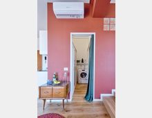 Interno domestico con parete color terracotta e condizionatore a parete sopra una porta che conduce a un piccolo locale lavanderia con lavatrice e mensole. In primo piano pavimento in legno, un mobiletto vintage con oggetti e una scala laterale con inserti in vetrocemento.