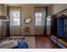 Interno di una camera da letto con due grandi finestre con tende leggere e drappeggi blu, pavimento in parquet e tappeto chiaro. Sono visibili una lampada da terra, una sedia decorata con specchio ovale a parete, un appendiabiti a vista e una panca imbottita.