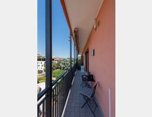 Balcone lungo e stretto di un appartamento con pavimento piastrellato, ringhiera metallica scura e parete esterna color pesca con porta finestra. In fondo si vede un piccolo tavolo con sedie e, oltre la ringhiera, una vista su alberi e palazzi sotto un cielo sereno.