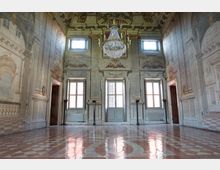 Interno di una sala storica decorata in stile barocco, con pareti affrescate raffiguranti colonne e architetture illusionistiche. La stanza presenta un pavimento in marmo a motivi geometrici, grandi finestre con cornici elaborate, e un lampadario monumentale al centro del soffitto decorato.